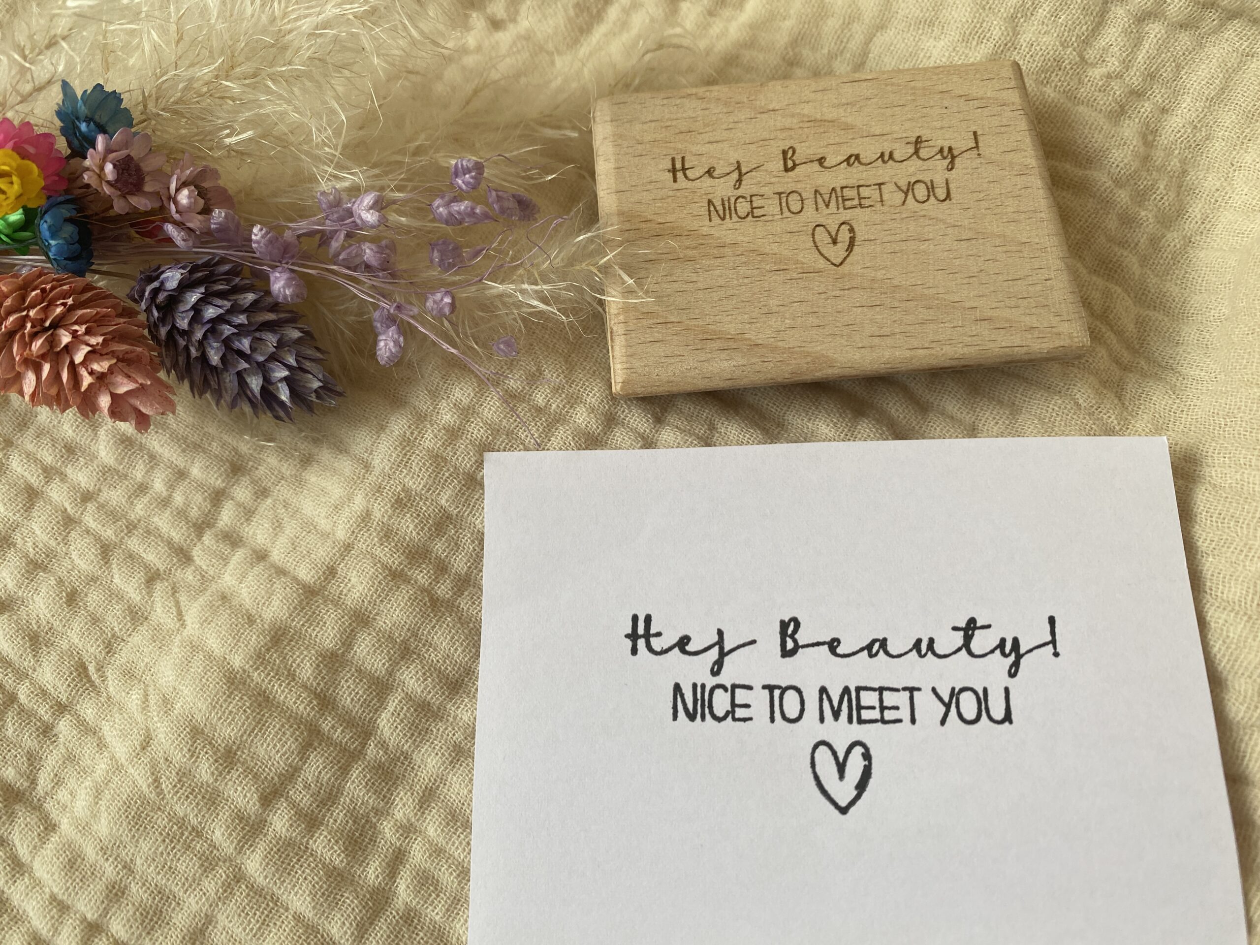 Das Bild zeigt einen rechteckigen Holzstempel, auf dem die Worte „Hey Beauty! NICE TO MEET YOU“ eingraviert sind, gefolgt von einem kleinen Herzsymbol. Der Stempel liegt auf einem beige-gelben Stoffhintergrund neben getrockneten Blumen in verschiedenen Farben. Auf dem darunterliegenden Stück Papier ist der Abdruck des Stempels zu sehen, der die gleiche Botschaft in klaren, schwarzen Buchstaben wiedergibt. Die Szene vermittelt eine warme, freundliche Stimmung und eignet sich gut für den persönlichen oder geschäftlichen Gebrauch, um eine positive Botschaft zu übermitteln.